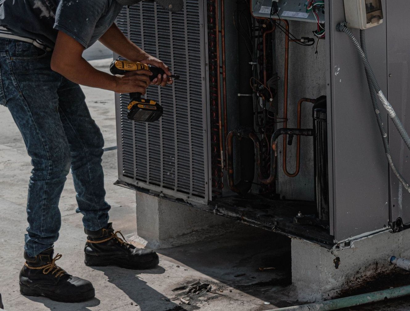 A worker in a bucket hat repairs an outdoor air conditioning unit on a rooftop.